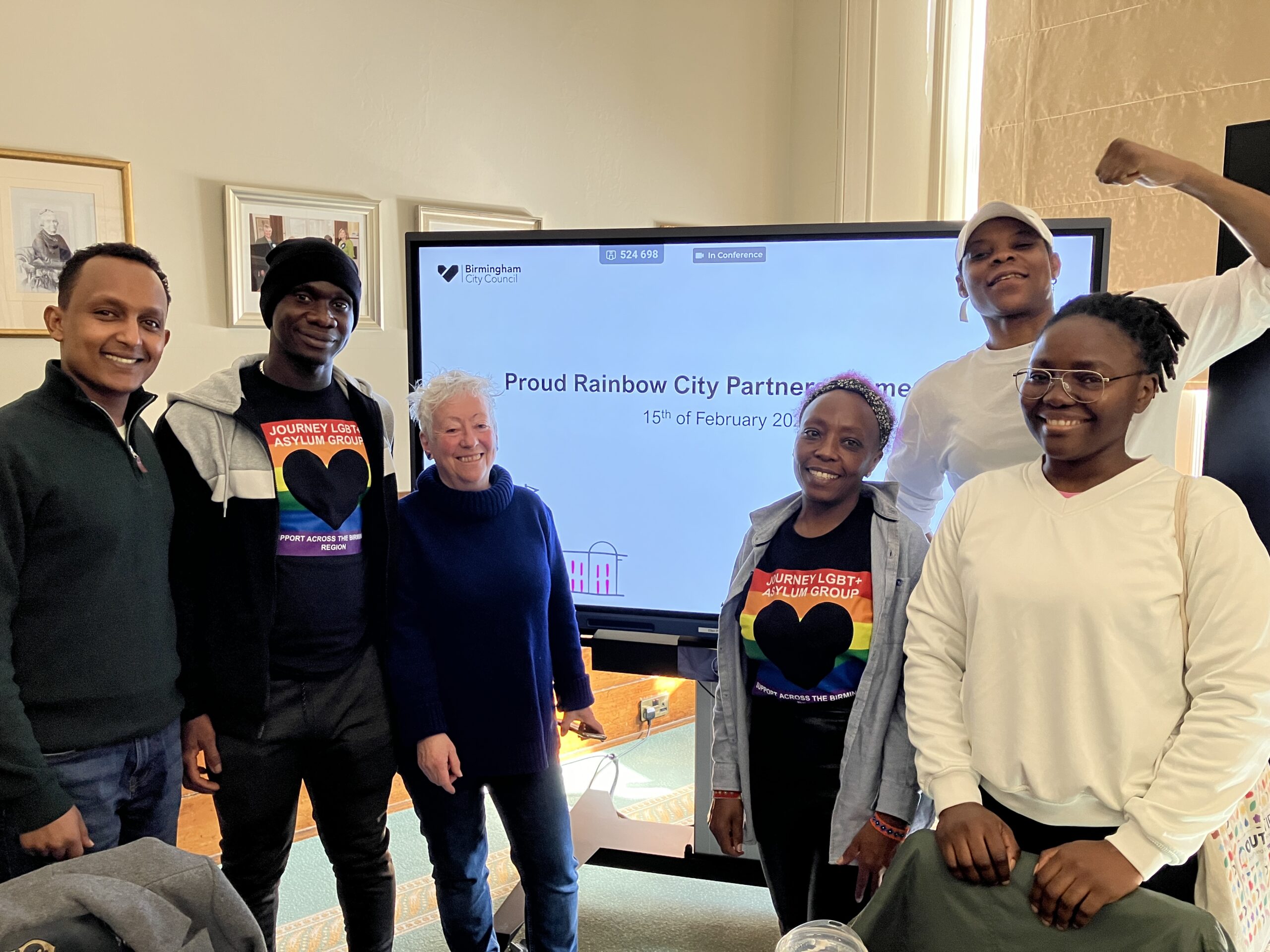 Five Journey members and Sheila stand smiling at the camera in front of a large screen displaying he text: Proud City Rainbow Partnership