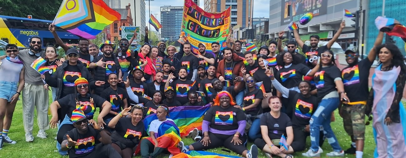 A large group of Journey members, many wearing Journey tshirts and waving Pride flags, gathered together and smiling